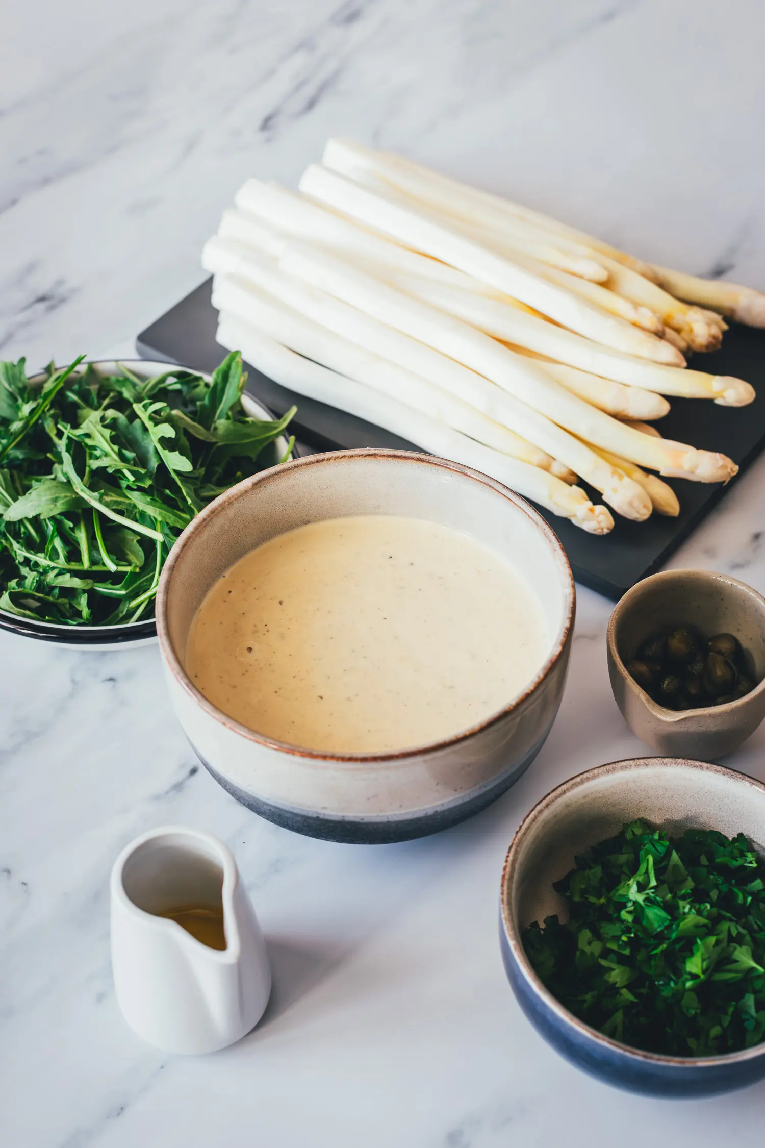 Cremige Tonnatosauce mit selbst hochgezogener Mayo in einer Keramikschale, daneben weißer Spargel, Rucola, Kapern und Olivenöl
