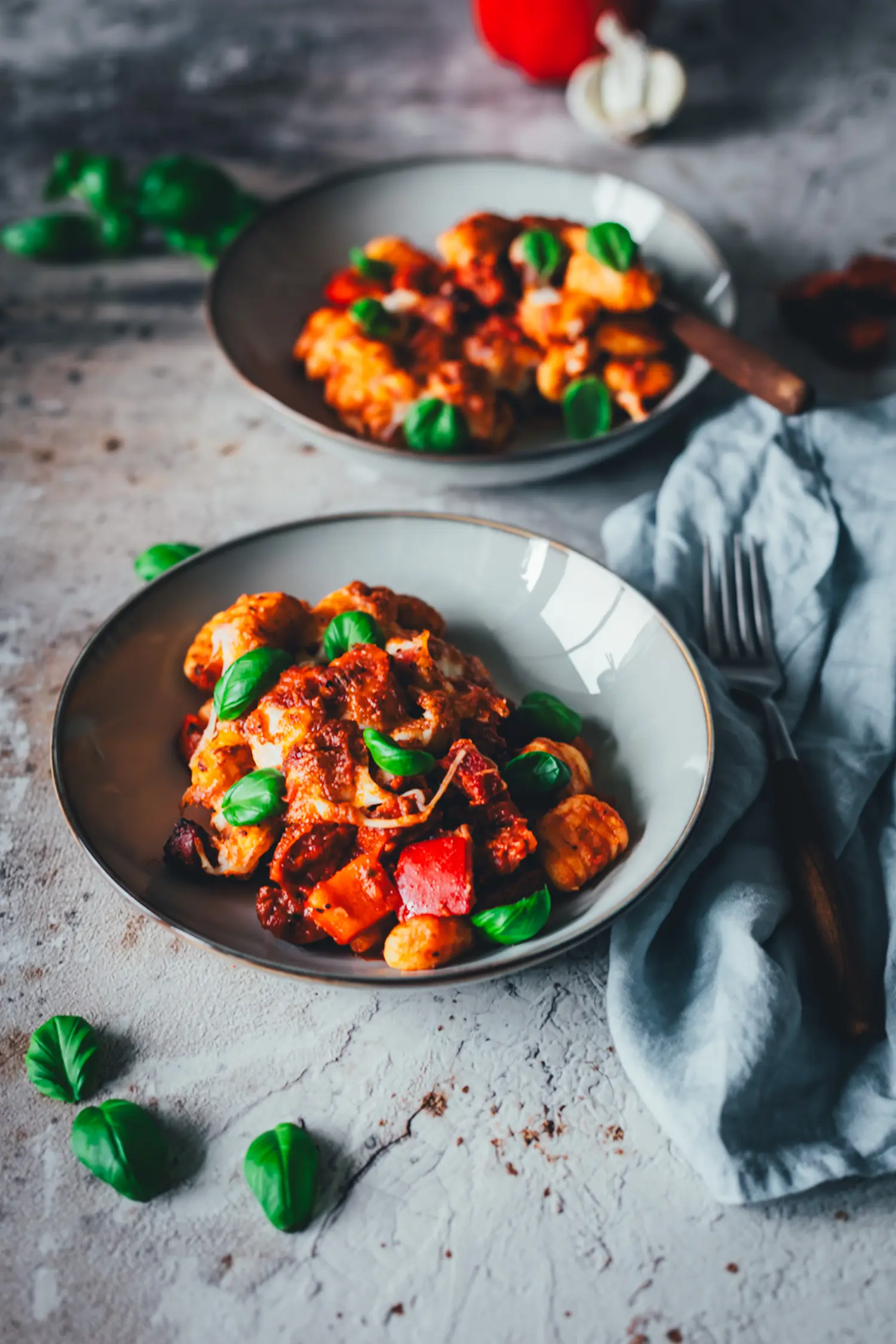 Gnocchi-Auflauf mit sichtbaren roten Paprikawürfeln und Chorizo in einer Schale, im Hintergrund ein zweiter Teller und frisches Basilikum