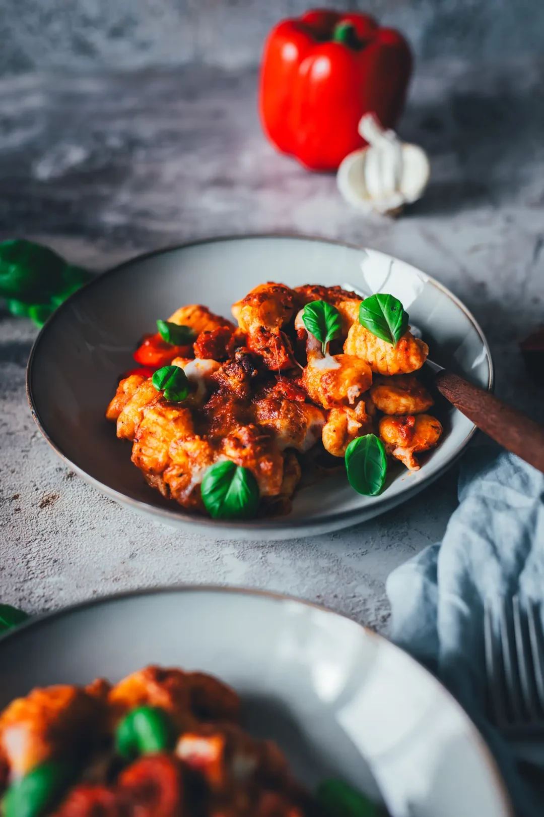 Teller mit Gnocchi-Auflauf aus dem Airfryer, überbacken mit Mozzarella, dazu Paprikawürfel, Chorizo und frische Basilikumblätter, im Hintergrund eine rote Paprikaschote und eine Knoblauchknolle