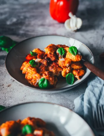Teller mit Gnocchi-Auflauf aus dem Airfryer, überbacken mit Mozzarella, dazu Paprikawürfel, Chorizo und frische Basilikumblätter, im Hintergrund eine rote Paprikaschote und eine Knoblauchknolle