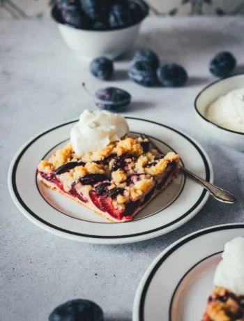 Ein weißer Teller mit einem Stück Zwetschgenkuchen mit Hefeteig und Streuseln, serviert mit Schlagsahne. Im Hintergrund violette Zwetschgen, im Vordergrund ein weiterer Teller.
