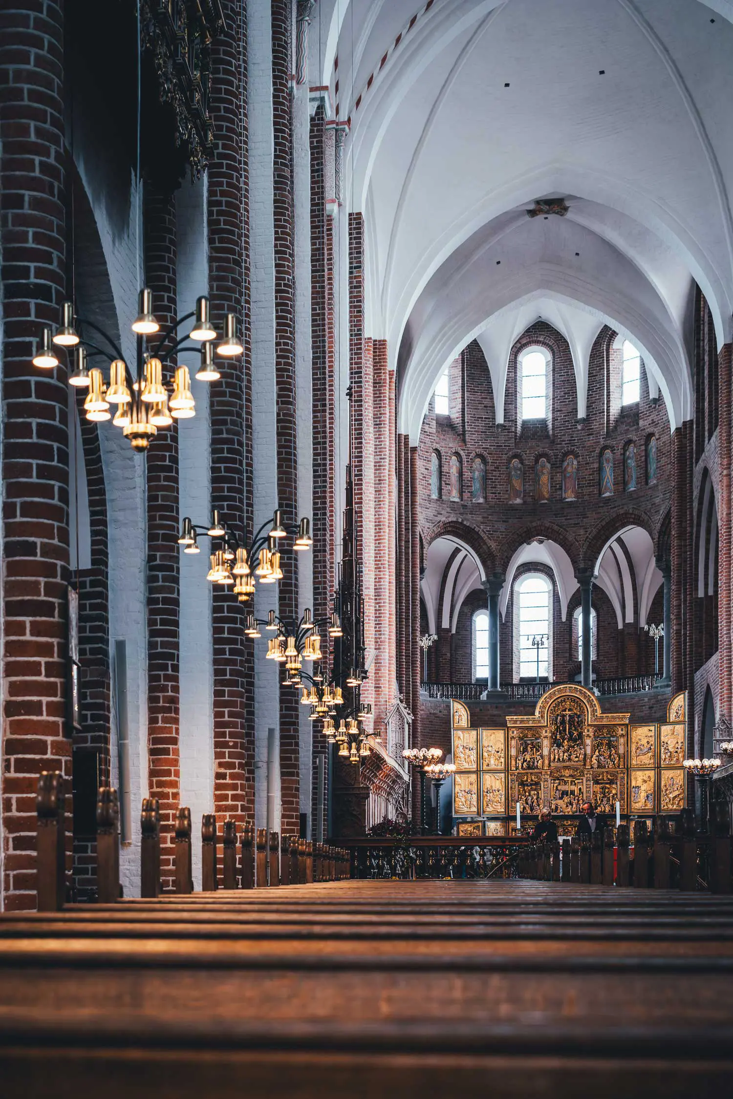Mittelschiff der Domkirke Roskilde mit Holzbänken, Backsteinpfeilern, weißem Gewölbe, modernen Messingleuchtern und dem goldenen Altar im Chor