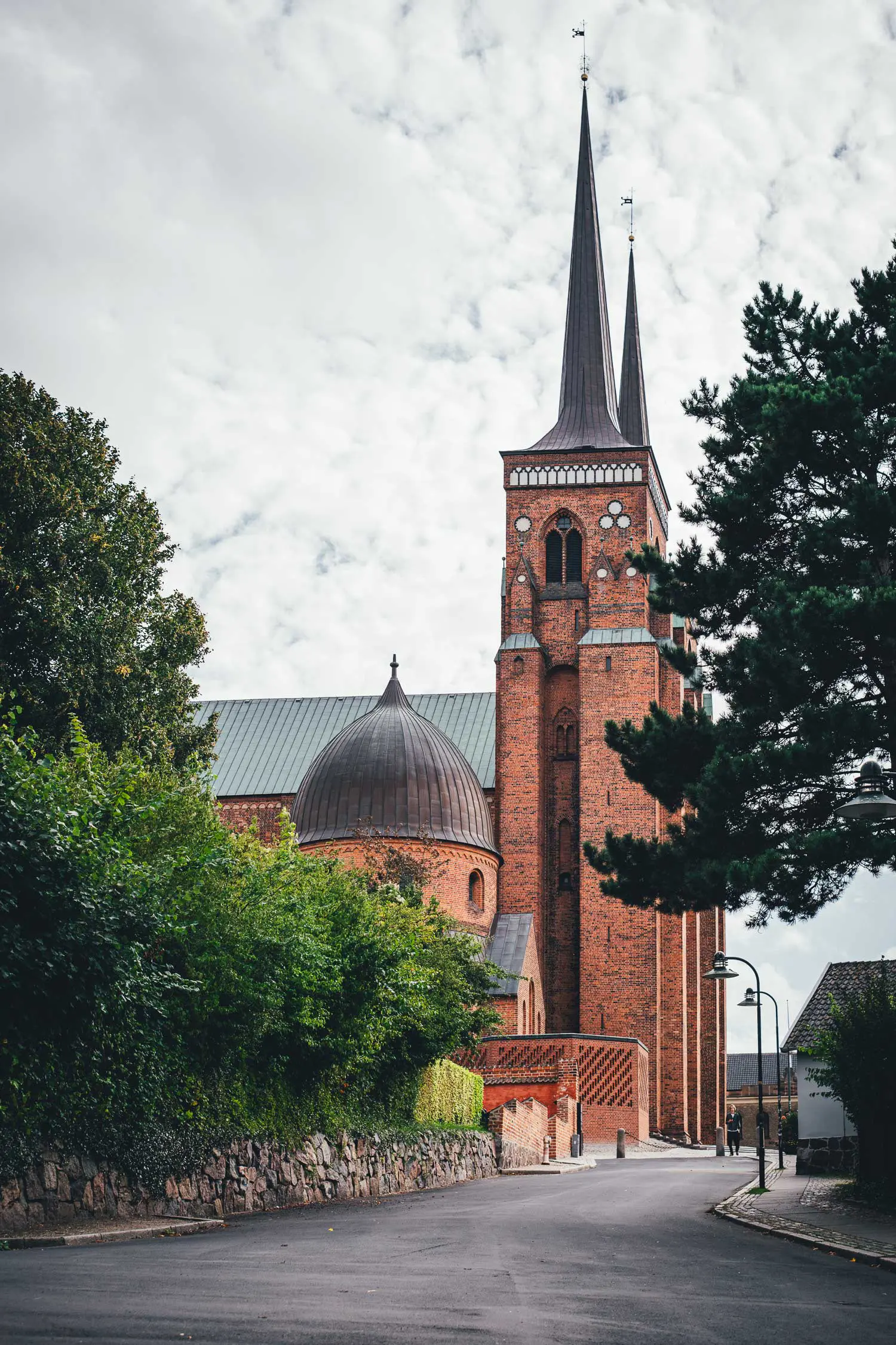 Blick von einer Straße in Roskilde auf die Backsteinkirche Domkirke mit zwei spitzen Türmen und einer Kupferkuppel, links Bäume und eine Natursteinmauer