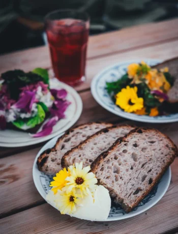 Teller mit Sauerteigbrot und Butter, dahinter zwei Smørrebrød mit essbaren Blüten auf einem Holztisch im Gewächshaus der Sct. Hans Have in Roskilde