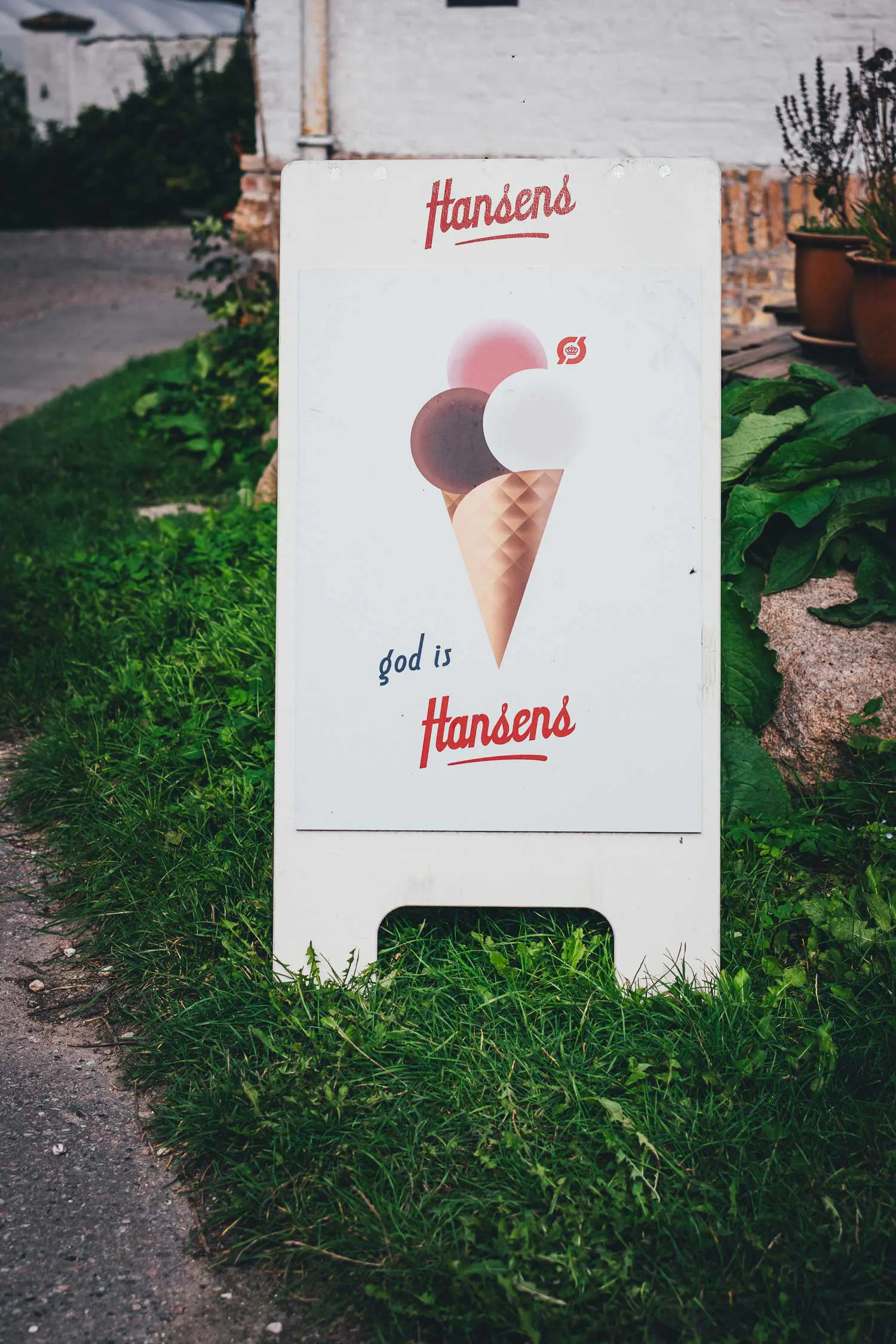 Weißer Aufsteller mit dem Logo Hansens, einer gezeichneten Eiswaffel mit drei Kugeln und dem Schriftzug god is Hansens auf einer Wiese vor dem Café von Svanholm