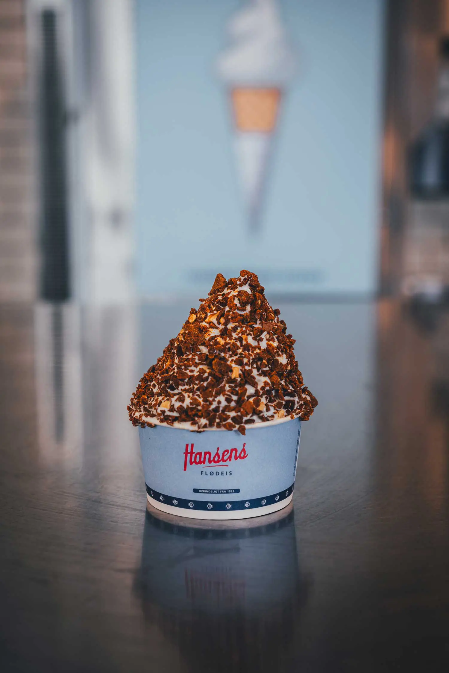 Hoch gezapftes Sahneeis mit knusprigem Nougat-Topping in einem hellblauen Becher mit Hansens-Logo auf einer Metalltheke bei Hansens Is in Jægerspris, im Hintergrund unscharf ein Poster mit Softeis