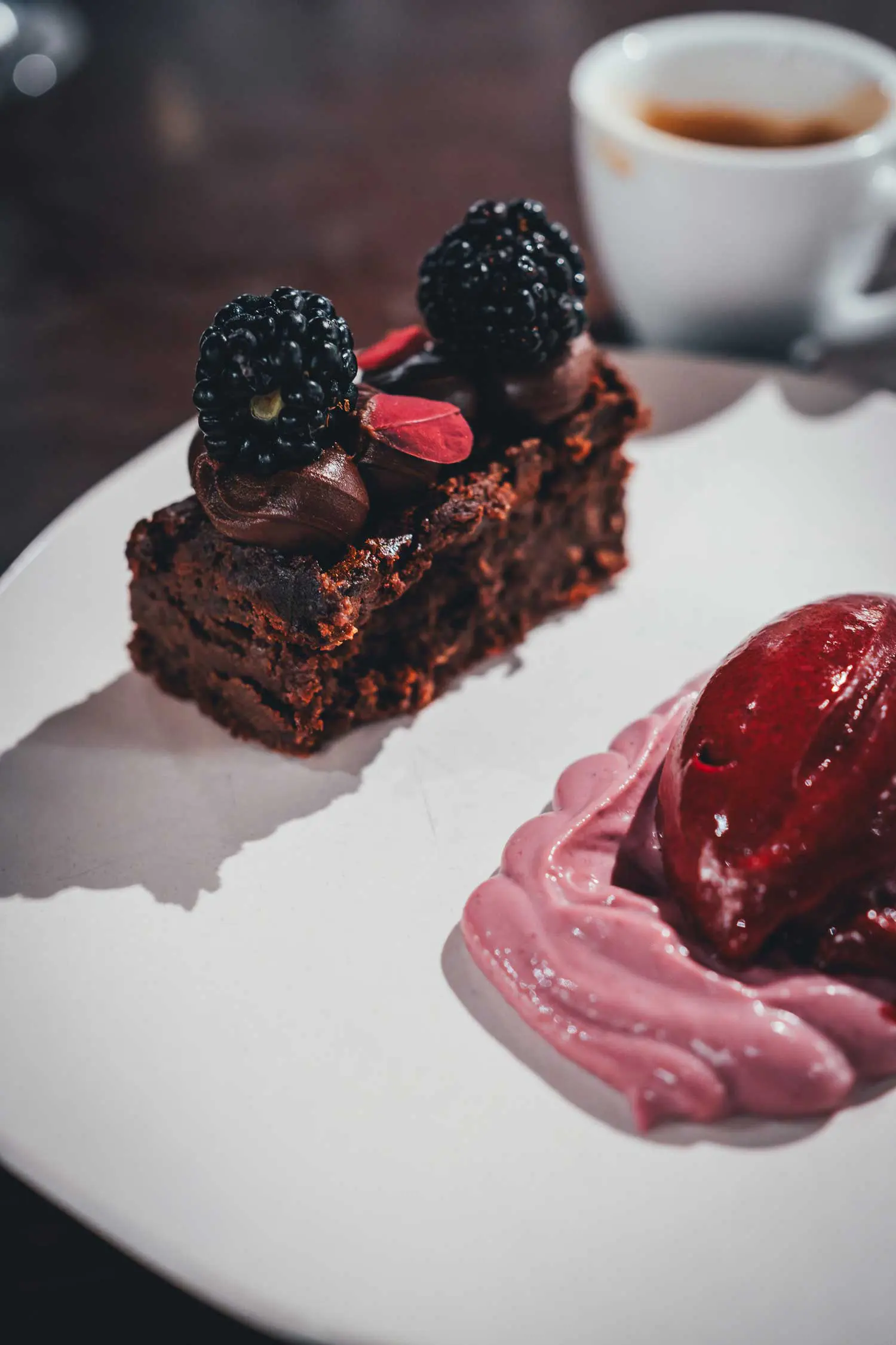 Stück Gâteau Marcel mit Schokoladencreme und frischen Brombeeren auf weißem Teller, daneben Brombeersorbet und ein Espresso
