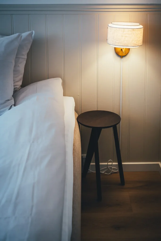 Frisch bezogenes Bett mit weißer Decke und Leinenkissen vor heller Holzvertäfelung, daneben ein Hocker als Nachttisch und eine Wandleuchte