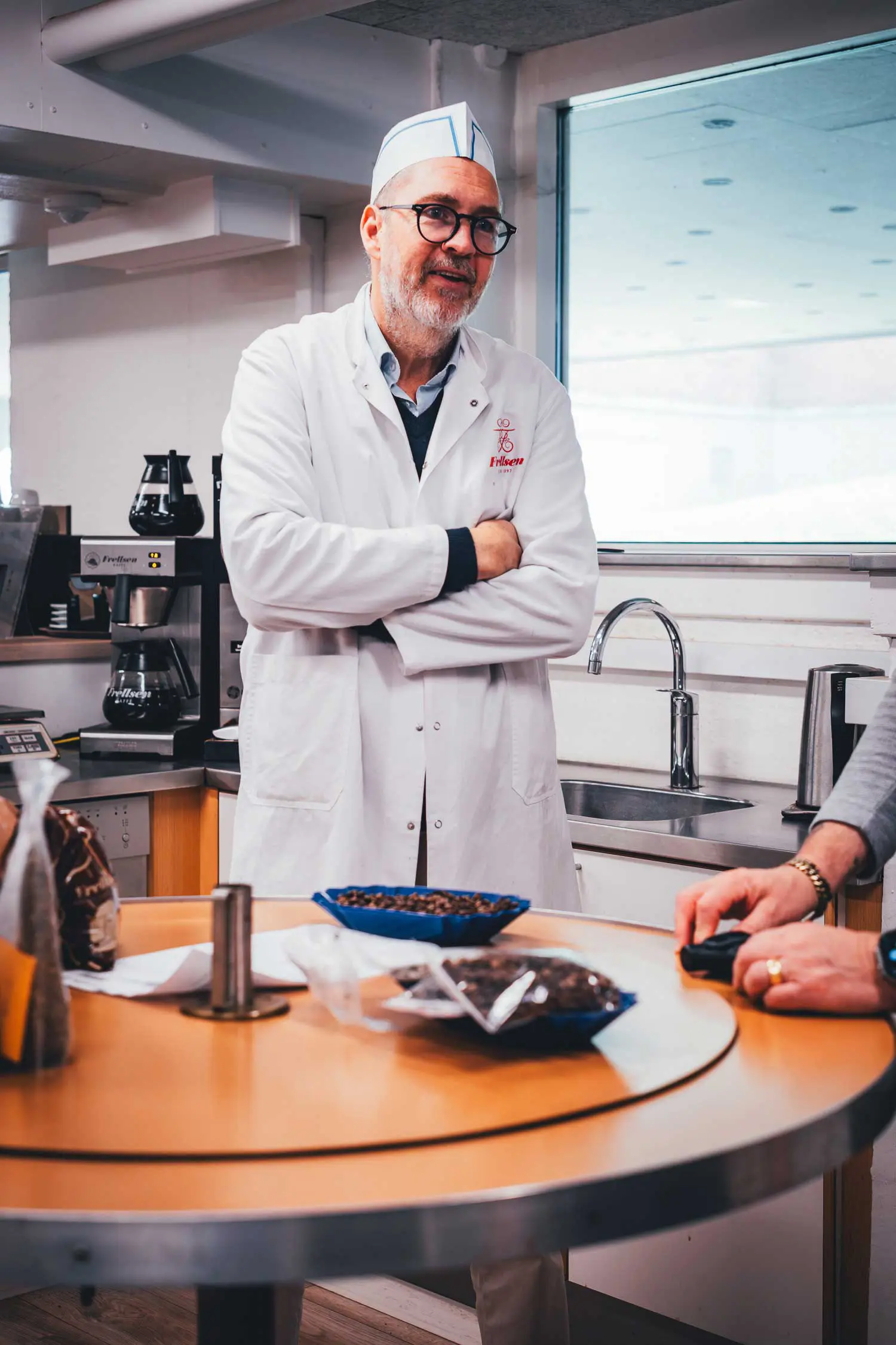 Peter Frellsen im weißen Kittel mit Firmenlogo steht vor einer Kaffeemaschine, auf dem Tisch Schalen mit Kaffeebohnen