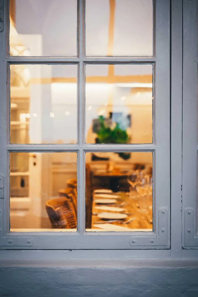 Blick durch ein weißes Sprossenfenster in den warm beleuchteten Gastraum des Pipers Hus in Roskilde mit gedeckter Tafel und Stühlen