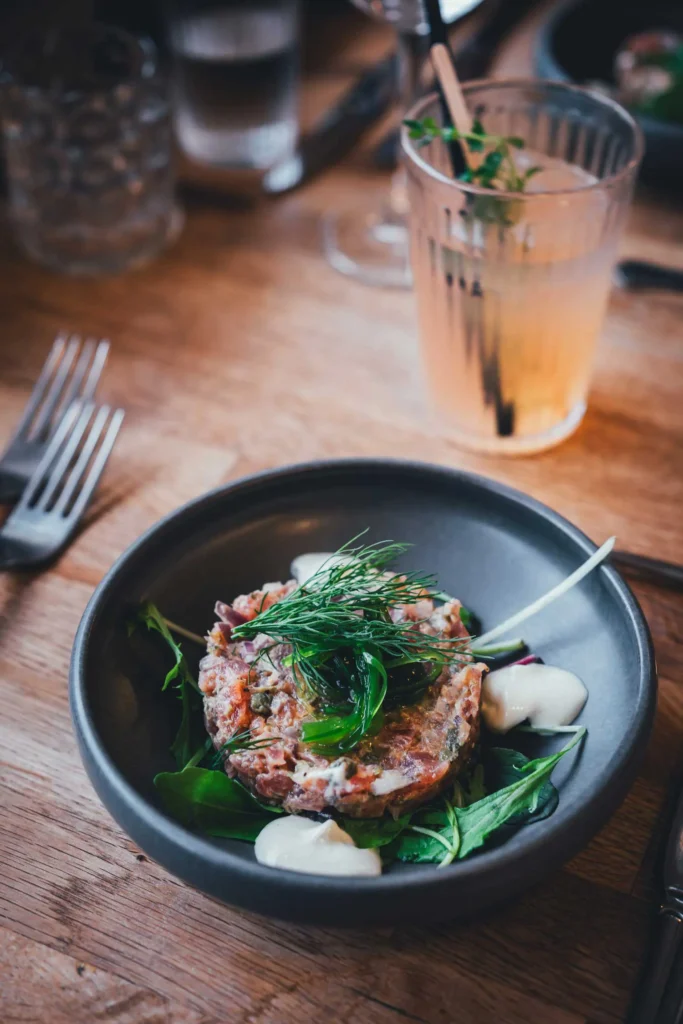 Tatar mit Dill, roten Zwiebeln und Klecksen heller Creme auf Blattsalat in einer dunklen Schale, dahinter ein Glas mit rosa Getränk, im Pipers Hus in Roskilde