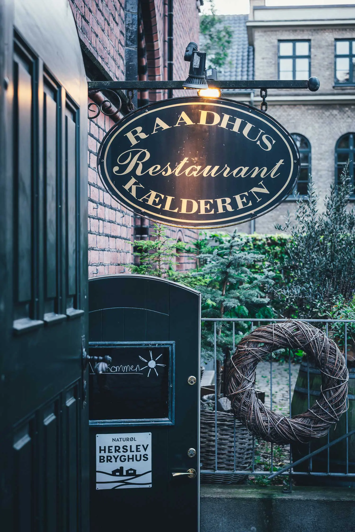 Schwarzes ovales Schild mit goldener Schrift Raadhus Restaurant Kælderen über einer dunkelgrünen Tür in Roskilde, an der Tür eine Plakette von Herslev Bryghus, daneben ein Weidenkranz am Zaun