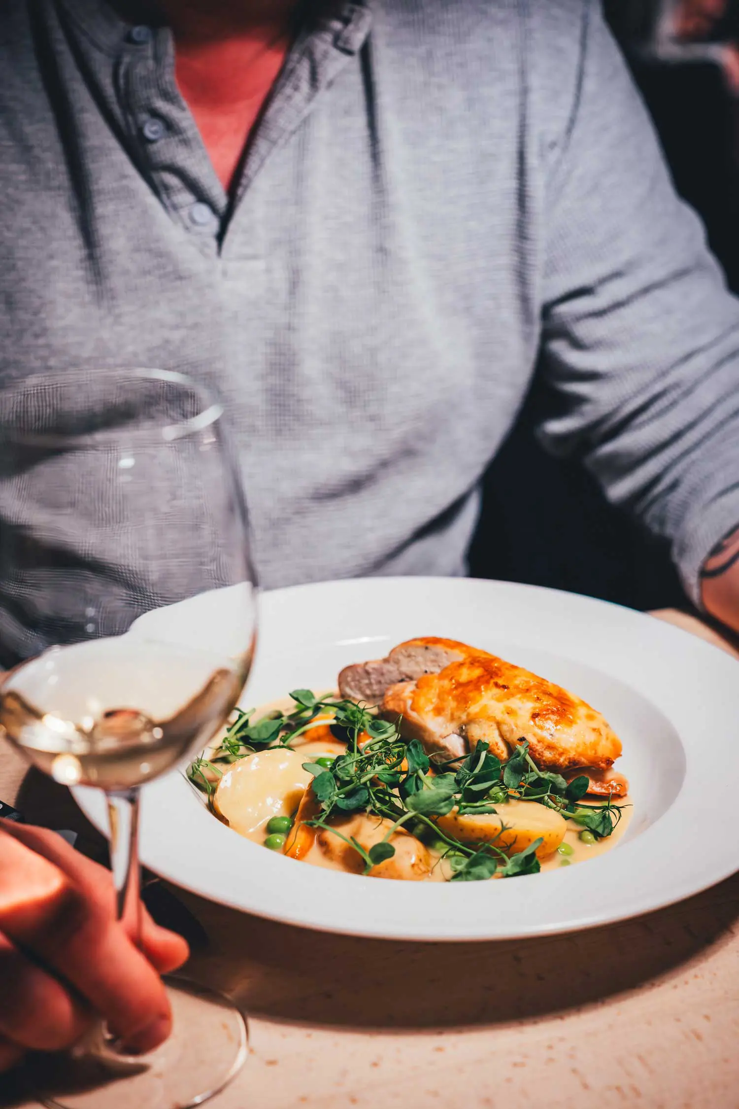 Tiefer weißer Teller mit gebratenem Hühnchen, Kartoffeln, Erbsen und Erbsensprossen in heller Sauce im Raadhuskælderen Roskilde, davor ein Glas Weißwein