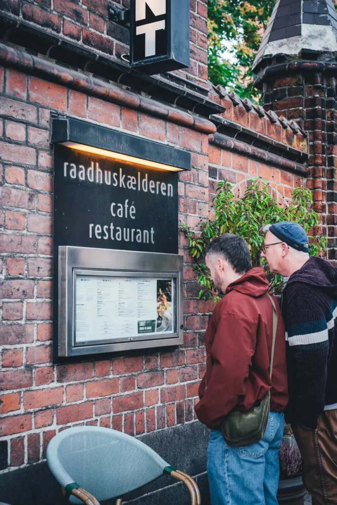 Sascha und Torsten von Die Jungs kochen und backen lesen die Speisekarte im Schaukasten an der Backsteinmauer des Raadhuskælderen in Roskilde