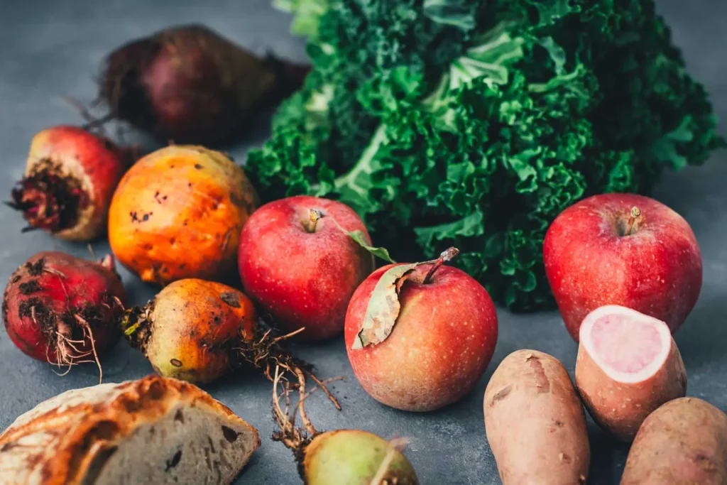 Rote und gelbe Bete mit Wurzeln, ein Bund Grünkohl, rote Äpfel, rote Kartoffeln, eine davon aufgeschnitten, und ein Stück Brot auf einem dunkelgrauen Untergrund.