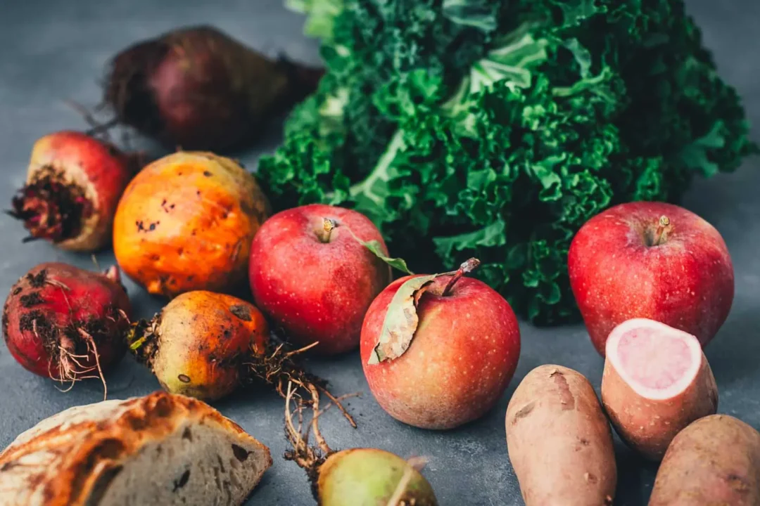Rote und gelbe Bete mit Wurzeln, ein Bund Grünkohl, rote Äpfel, rote Kartoffeln, eine davon aufgeschnitten, und ein Stück Brot auf einem dunkelgrauen Untergrund.