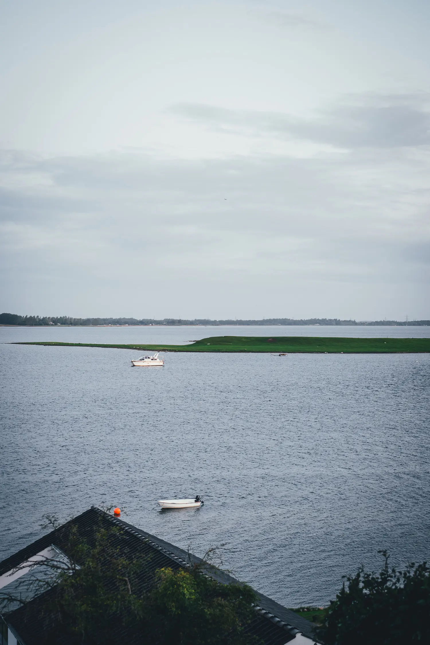 Blick vom Hotel Søfryd über ein dunkles Dach auf den Roskilde Fjord mit zwei kleinen Booten und einer grünen Landzunge unter grauem Himmel
