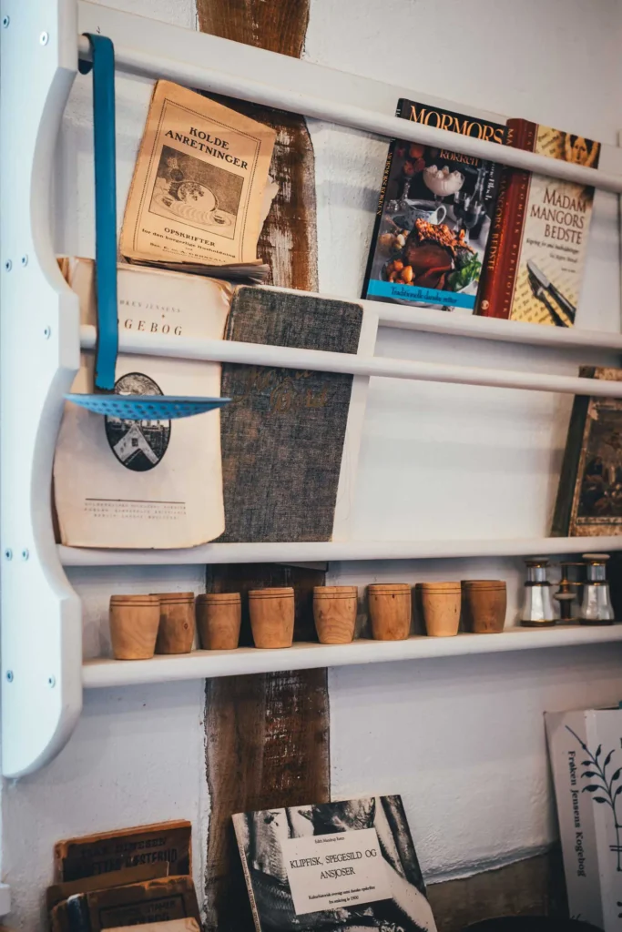 Weißes Wandregal mit alten dänischen Kochbüchern und Holzbechern im Café Hvidesøhus in Sagnlandet Lejre