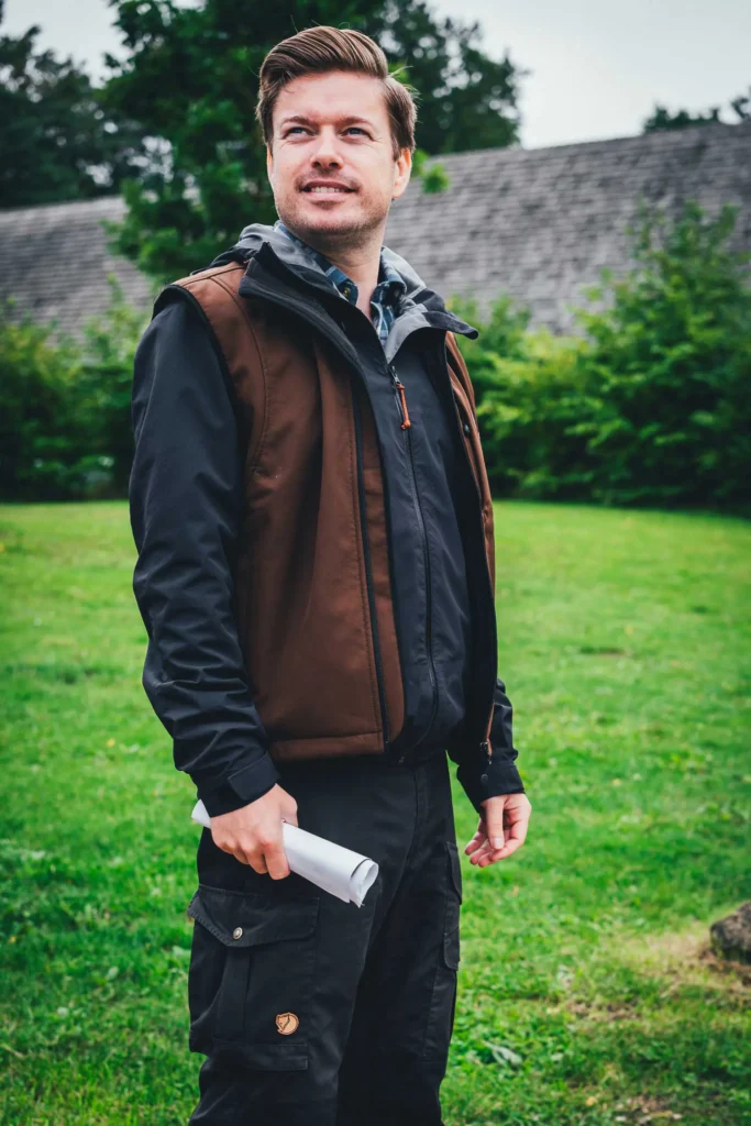 Kenneth, Guide in Sagnlandet Lejre, in brauner Weste mit einem zusammengerollten Blatt in der Hand vor einem reetgedeckten Haus