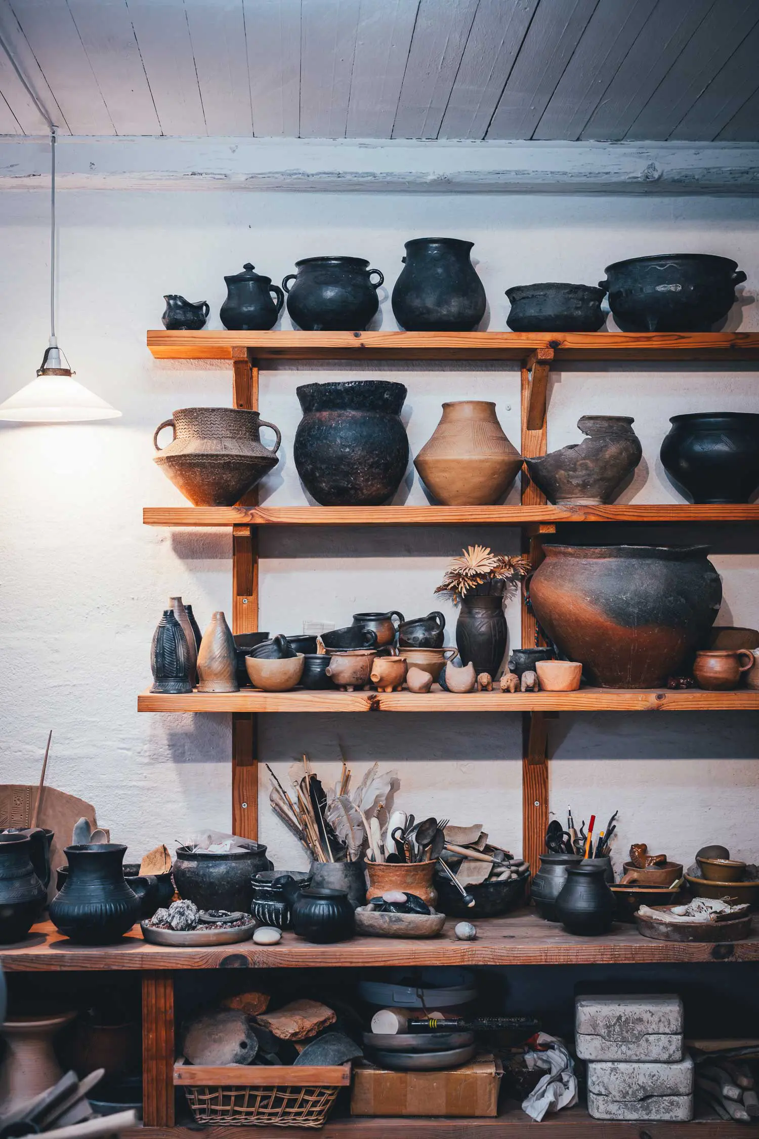 Holzregal in der Töpferei von Sagnlandet Lejre mit schwarz gebrannten Jydepotter und weiteren Tontöpfen in verschiedenen Größen, darunter kleine Gefäße und Werkzeug