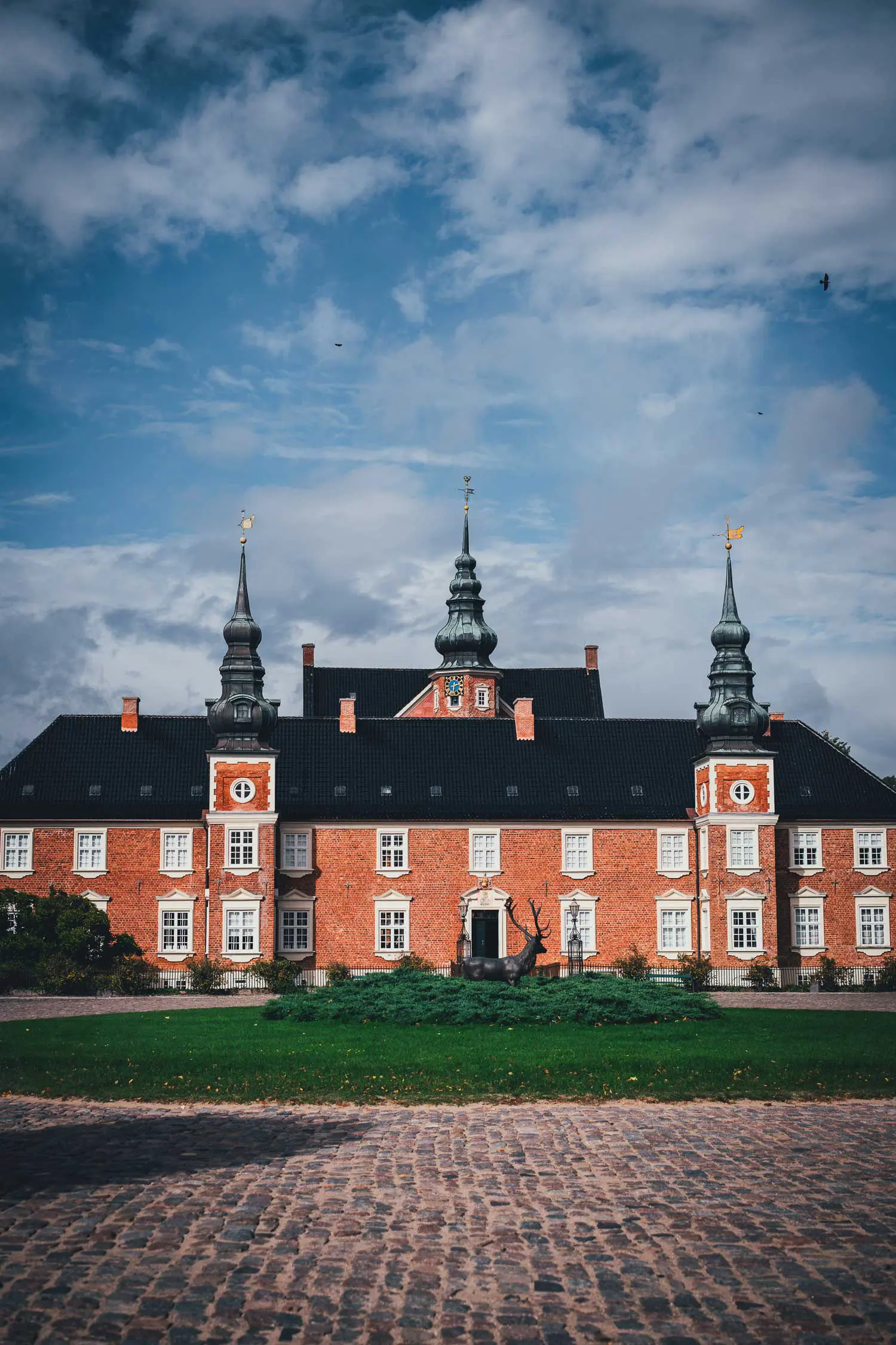 Schloss Jægerspris aus rotem Backstein mit drei schwarzen Zwiebeltürmen und weißen Fensterrahmen, davor eine Rasenfläche mit Hirschskulptur und Kopfsteinpflaster unter bewölktem Himmel