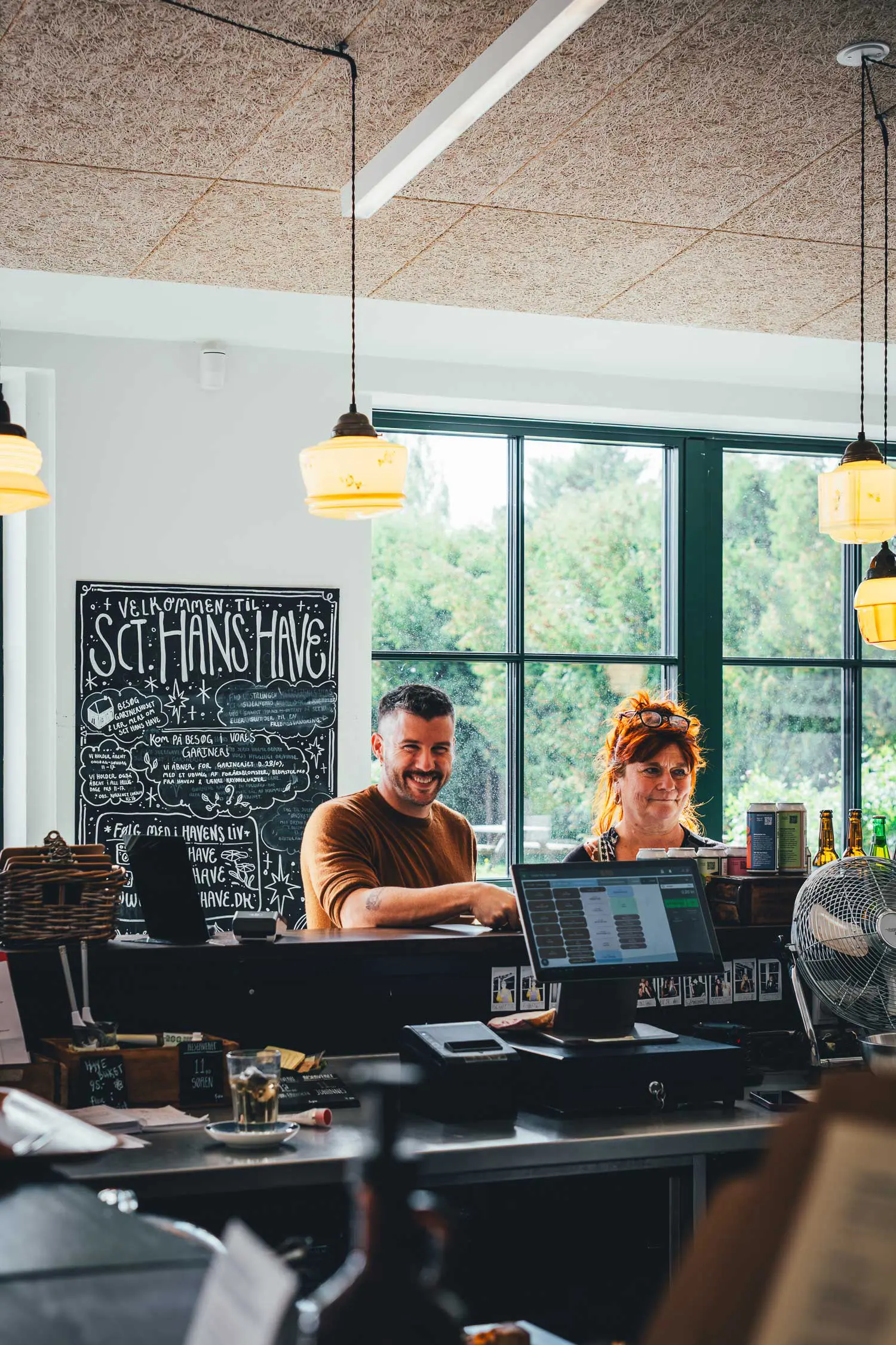 Zwei Personen stehen lachend hinter der Theke des Cafés, dahinter eine beschriebene Tafel mit dem Willkommensgruß und ein großes Fenster