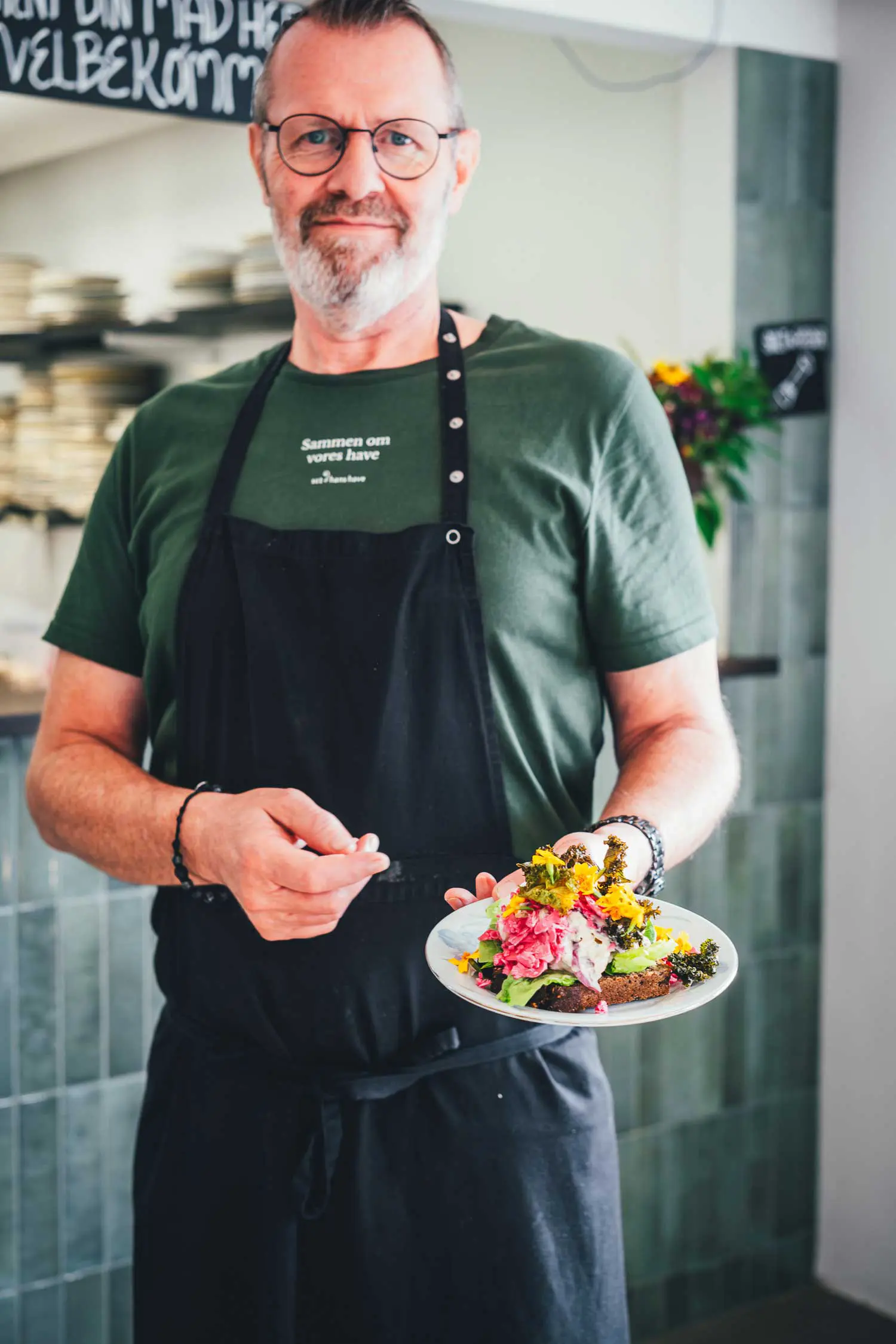 Mann mit Brille und schwarzer Schürze hält ein Smørrebrød mit Blüten und Grünkohlchips auf einem Teller in die Kamera