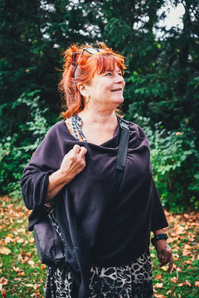 Frau mit roten Haaren und Brille im Haar steht vor dunklen Bäumen und schaut lächelnd zur Seite