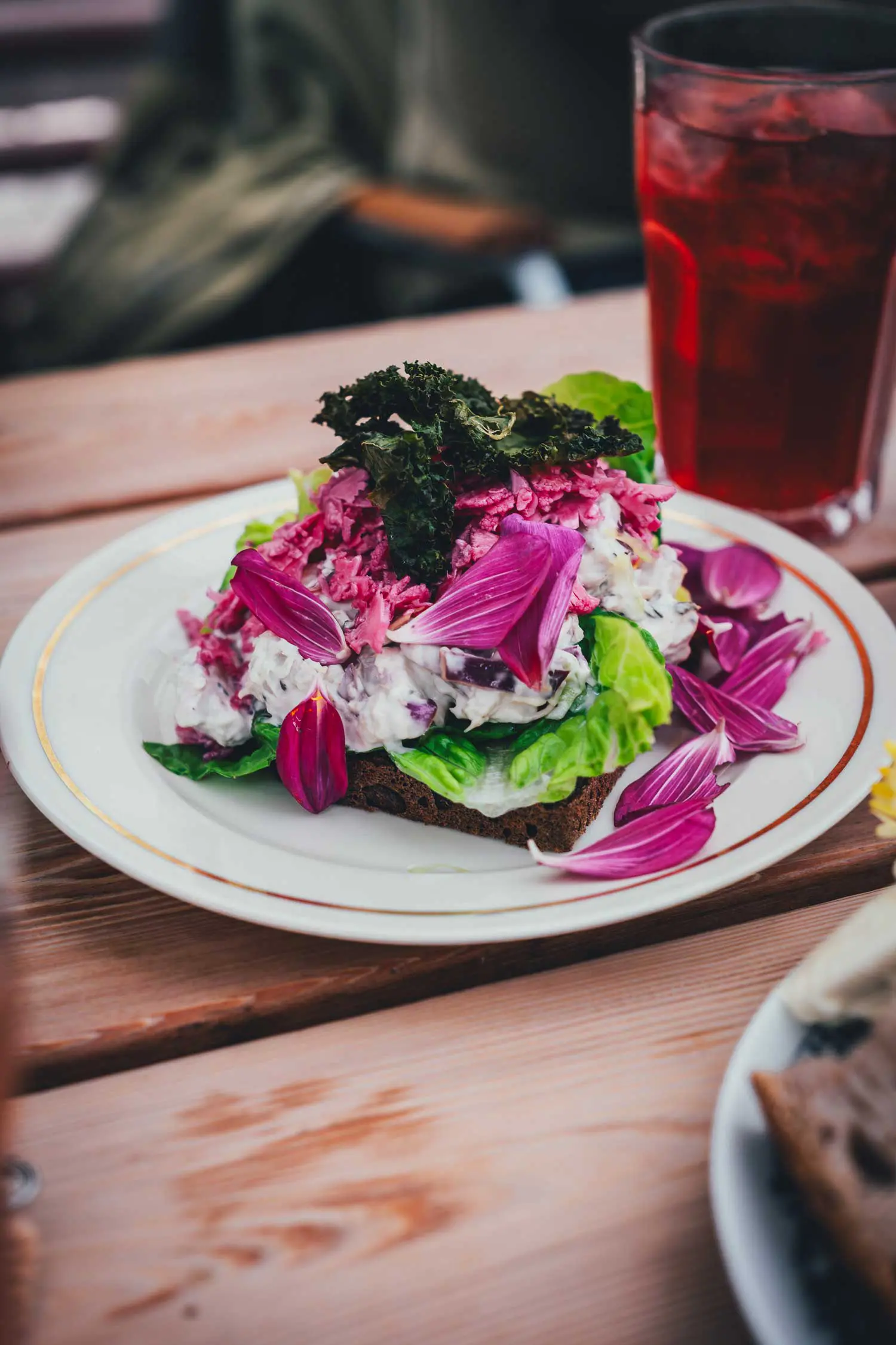 Smørrebrød auf dunklem Roggenbrot mit Salat, eingelegtem Rotkohl und pinkfarbenen Blütenblättern, daneben ein Glas mit roter Limonade