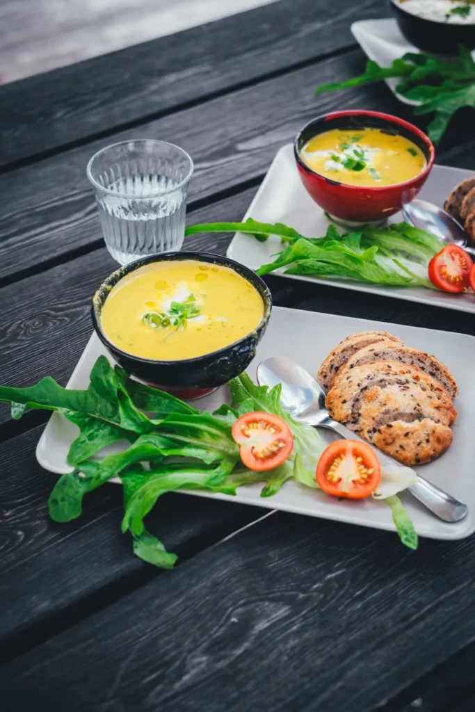 Zwei Schalen gelbe Gemüsesuppe mit Frühlingszwiebeln und einem Klecks Creme, dazu Körnerbrot, Blattsalat und halbierte Tomaten auf hellen Tellern auf einem schwarzen Holztisch im Café von Svanholm