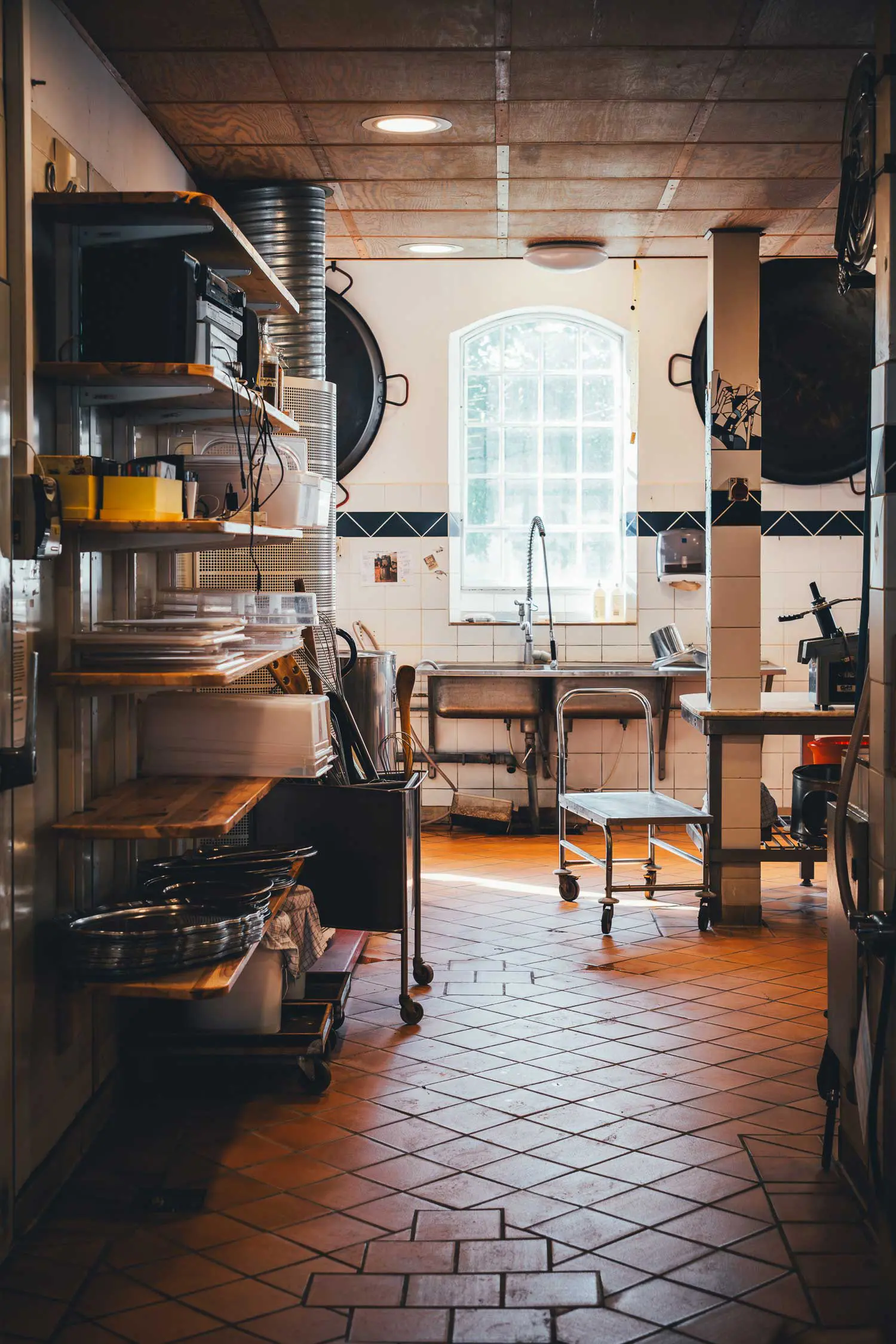 Großküche auf Svanholm mit Edelstahlspüle unter einem Rundbogenfenster, Regalen mit Blechen und Schüsseln, Rollwagen und rotem Fliesenboden