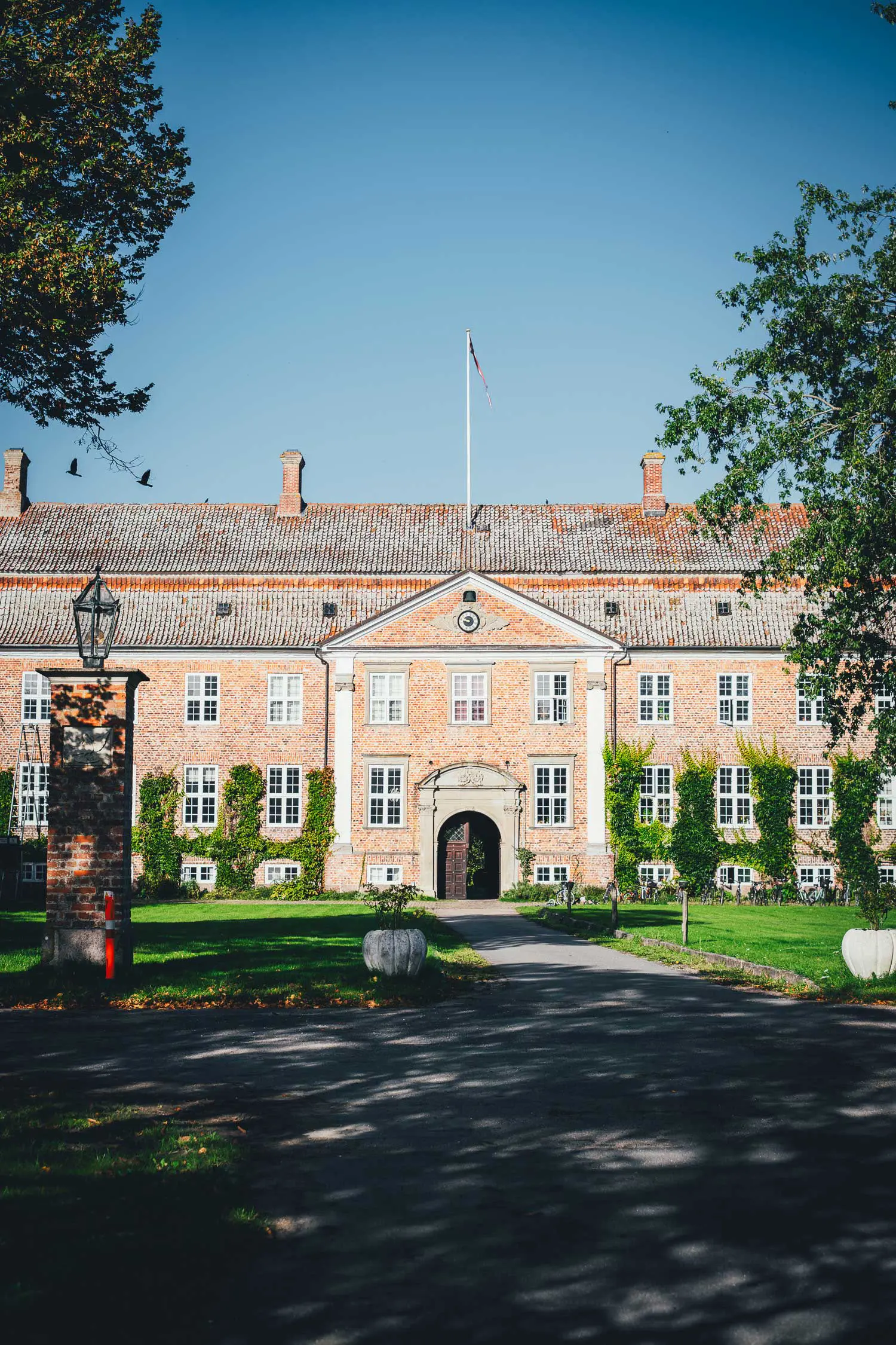 Das Haupthaus des Gutshofs Svanholm aus rotem Backstein mit weißen Sprossenfenstern, Giebel und Fahnenmast, davor eine Rasenfläche mit Weg zum Eingang