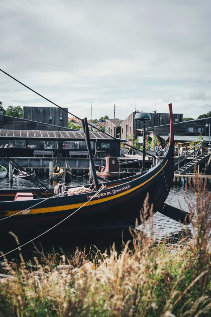 Schwarzer Wikingerschiff-Nachbau mit gelb-rotem Streifen und dem Namen Havhingsten fra Glendalough am Bug liegt im Hafen des Wikingerschiffmuseums Roskilde, dahinter die Holzgebäude des Museums